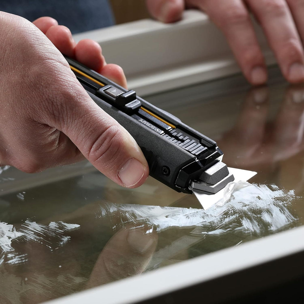 Close-up of a hand scraping off white paint residue from a glass window pane using the 2-in-1 utility scraper.