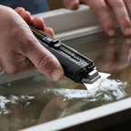 Close-up of a hand scraping off white paint residue from a glass window pane using the 2-in-1 utility scraper.