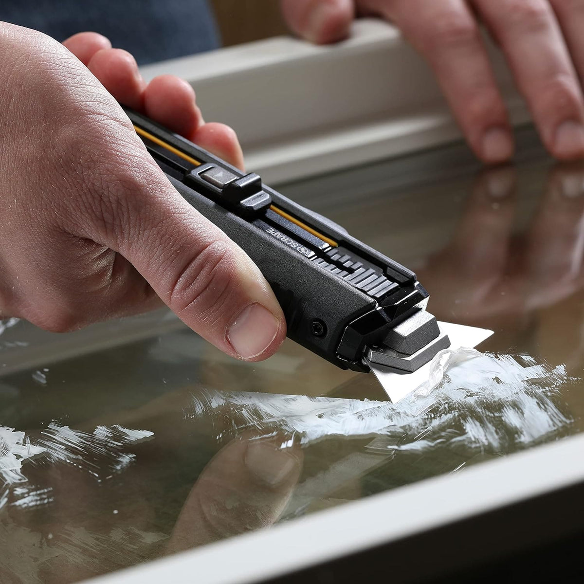 Close-up of a hand scraping off white paint residue from a glass window pane using the 2-in-1 utility scraper.