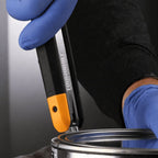 Gloved hand using the integrated can opener located at the base of the utility knife handle to open a paint can.