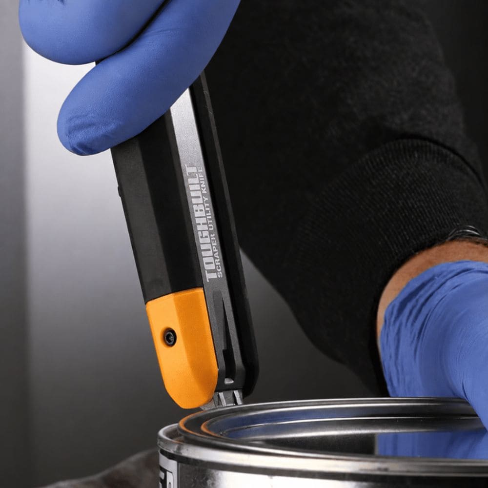Gloved hand using the integrated can opener located at the base of the utility knife handle to open a paint can.