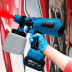 Gloved hand operating a blue cordless paint spray gun applying red paint against an outdoor wall.
