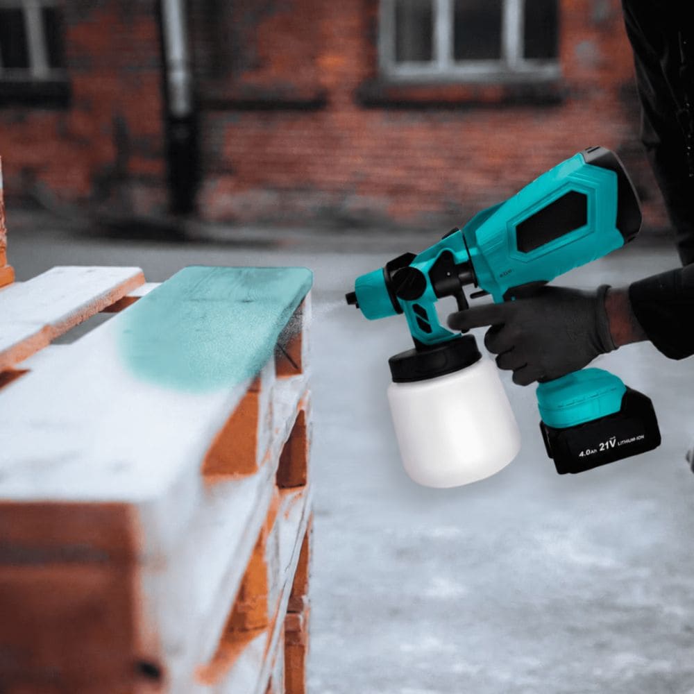 Person using a cordless paint gun to spray turquoise paint onto a wooden pallet outdoors.