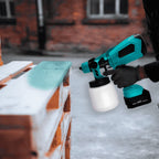 Person using a cordless paint gun to spray turquoise paint onto a wooden pallet outdoors.
